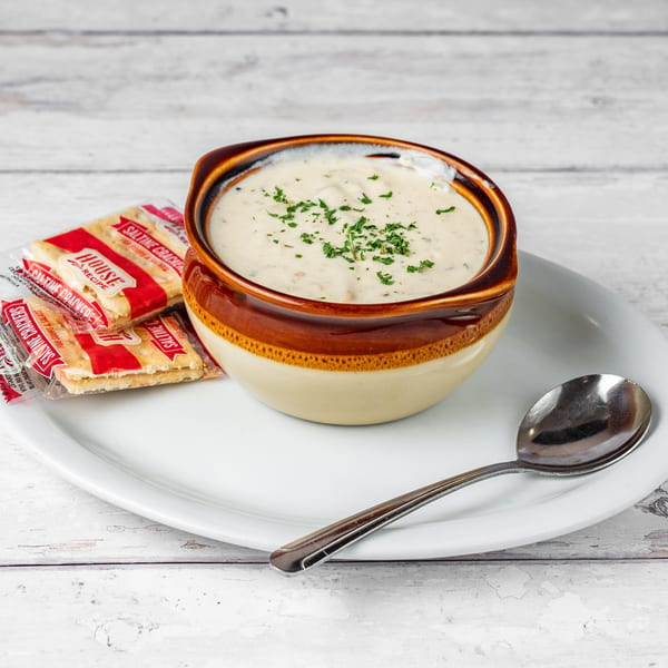 Creamy clam chowder in a brown and beige bowl, garnished with parsley, served with saltine crackers and a spoon.