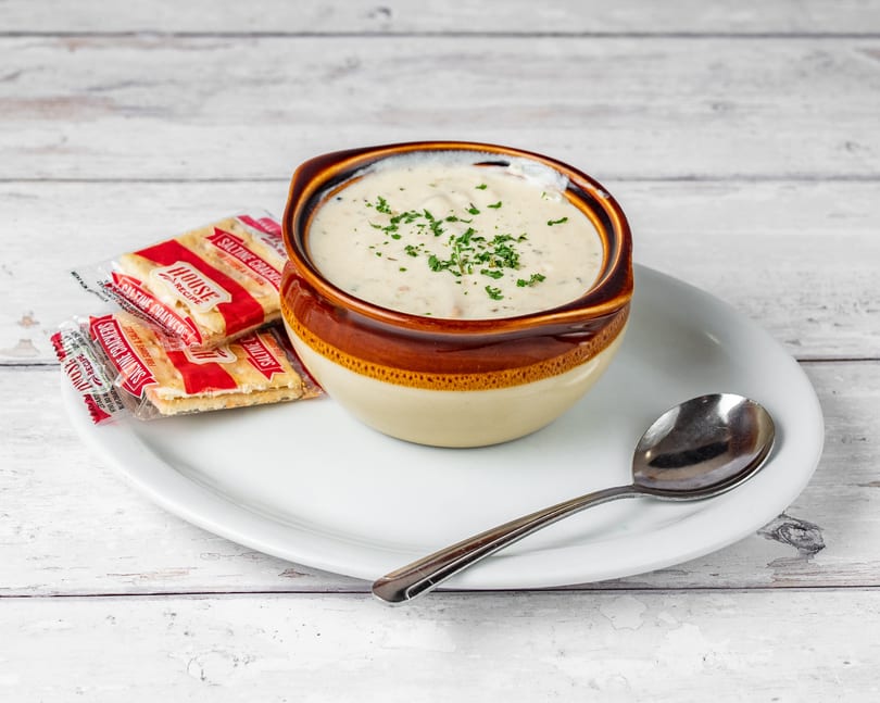 Creamy clam chowder in a brown and beige bowl, garnished with parsley, served with saltine crackers and a spoon.