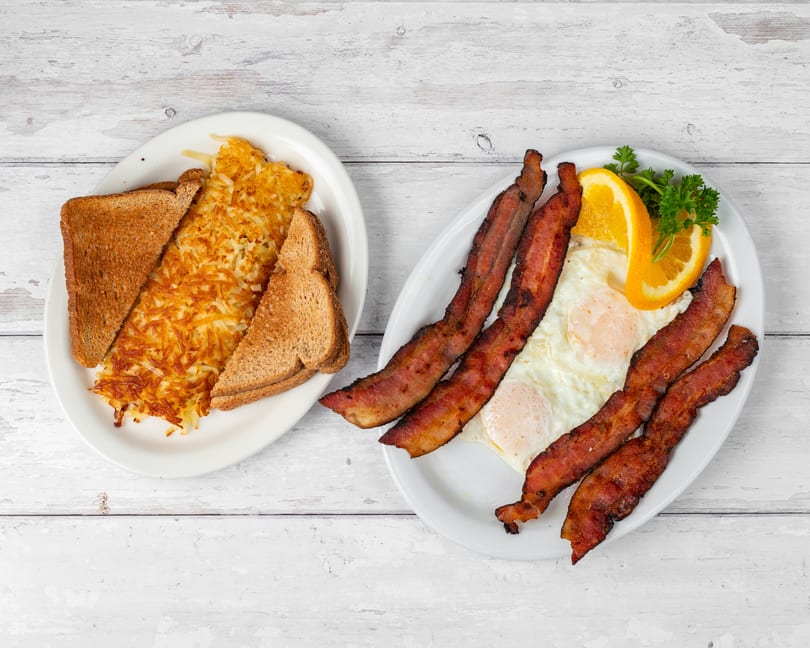 Two plates of breakfast food, one with hash browns and toast, the other with bacon and eggs.