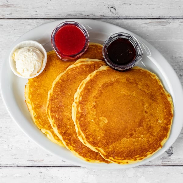 Three golden pancakes on a white plate with butter, strawberry jam, and maple syrup.